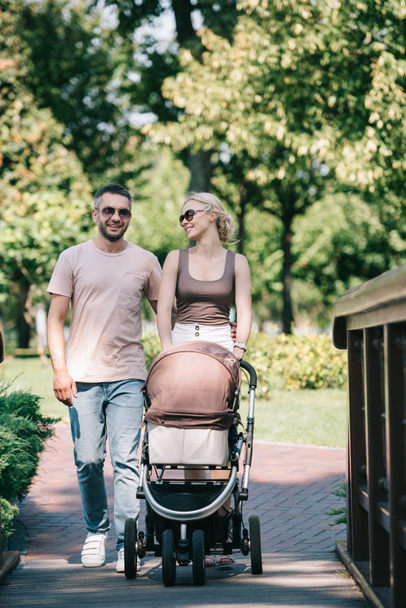 cheerful parents walking with baby carriage on bridge in park  - Photo, image