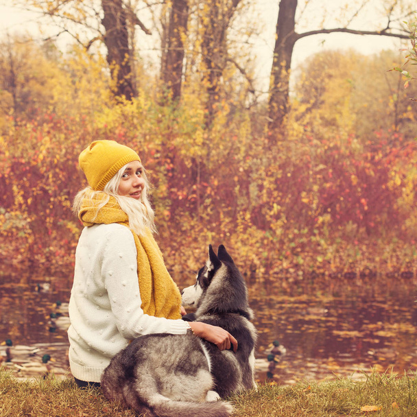 Jonge vrouw en haar hond zitten bij het meer buitenshuis in de herfst park  - Foto, afbeelding