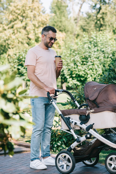 father walking with baby carriage in park and holding disposable coffee cup - Photo, Image
