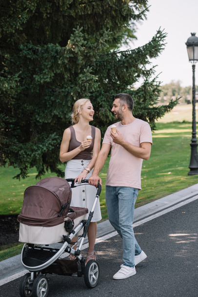 laughing parents walking with baby carriage and ice cream in park - Photo, Image
