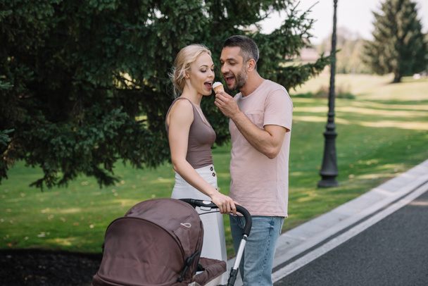 husband feeding wife with ice cream near baby carriage in park - Photo, Image