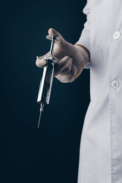 a doctor holding a big stainless steel syringe on dark background - Fotoğraf, Görsel
