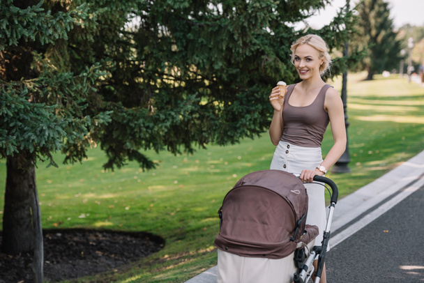 smiling mother walking with baby carriage and ice cream in park - Foto, imagen