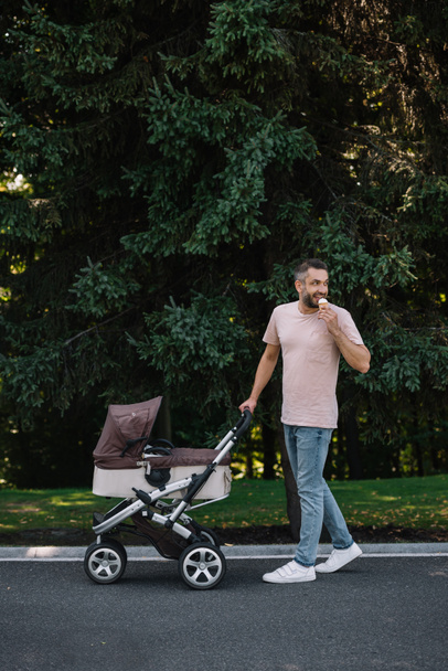 smiling father walking with baby carriage and ice cream in park and looking back - Zdjęcie, obraz