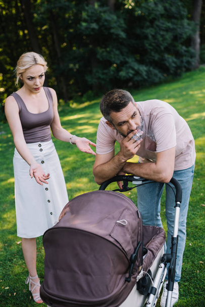 father smoking cigarette near baby carriage in park, angry mother gesturing and looking at him - Photo, Image