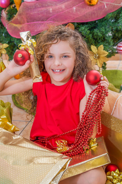 Close up of smiling girl wearing a red blouse and holding a christmas ball, decoration and gift in her hands, with a Christmas tree behind, christmas concept - Foto, immagini