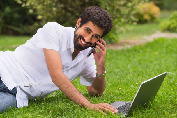 Młody szczęśliwy uśmiechający się mężczyzna pracujący z laptopa i telefon, na zewnątrz - Zdjęcie, obraz