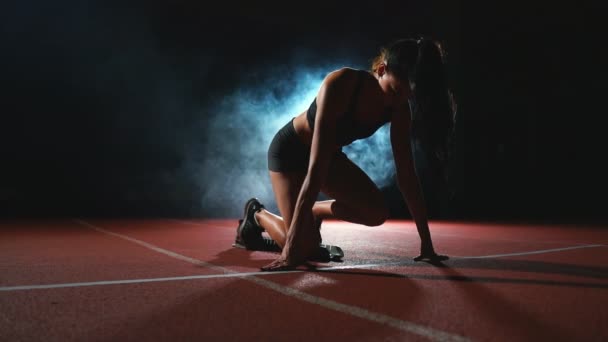 Nuori nainen urheilija mustissa shortseissa ja t-paita valmistautuu aloittamaan kilpailun 100 metrin juoksumatolla lähellä lähtöviivaa.
 - Materiaali, video