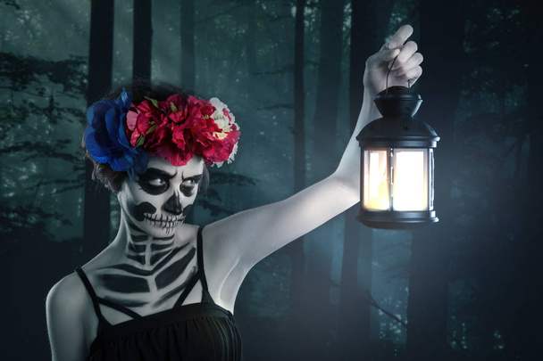 Bruja de Halloween. Hermosa mujer con máscara de Santa Muerte hechizo de lanzamiento sostiene un candelero contra el telón de fondo de un bosque de miedo
 - Foto, imagen