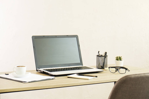 Laptop ohne Bildschirm, Tasse Cappuccino, Kaktus, Zubehör und zusammengefaltete Brille auf einem Holztisch in einem geräumigen Büro voller Sonnenlicht. kreativer Arbeitsbereich. Nahaufnahme, Kopierraum, Hintergrund - Foto, Bild