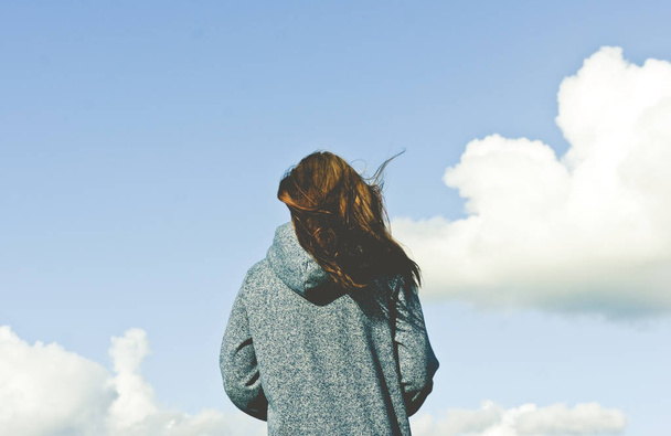the girl from the back goes against the blue sky with white clouds, is wearing a blue sweatshirt with a hood, the hair is developed by the wind, the sunny weather, the photo is up to the woman's belt - Photo, Image