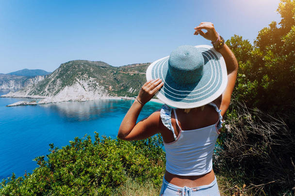 Petani beach Kefalonia. Young woman holding blue sun hat enjoying beautiful panorama of blue bay lagoon surrounded by steep cliff coastline. Greece - Photo, Image