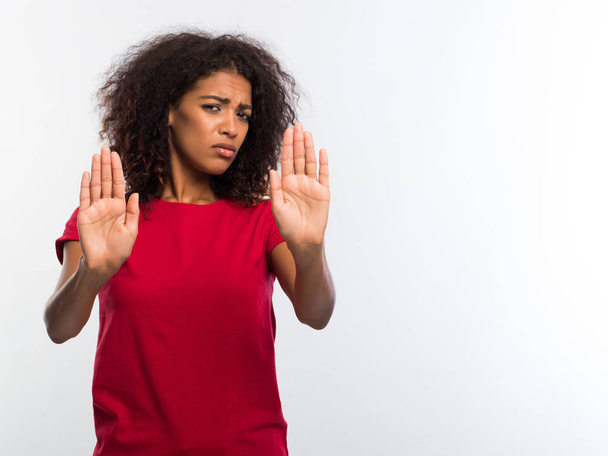 Portrait of serious young african woman in red t-shirt showing stop gesture with her palms isolated over white wall background. Decline concept. - Φωτογραφία, εικόνα