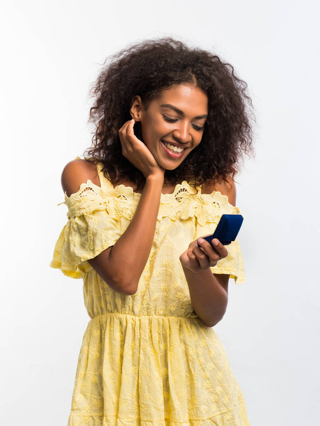 Счастливая молодая африканская девушка в желтом платье и соломенной шляпе с помощью смартфона на белом фоне. Технологическая концепция. Интернет-серфинг
. - Фото, изображение