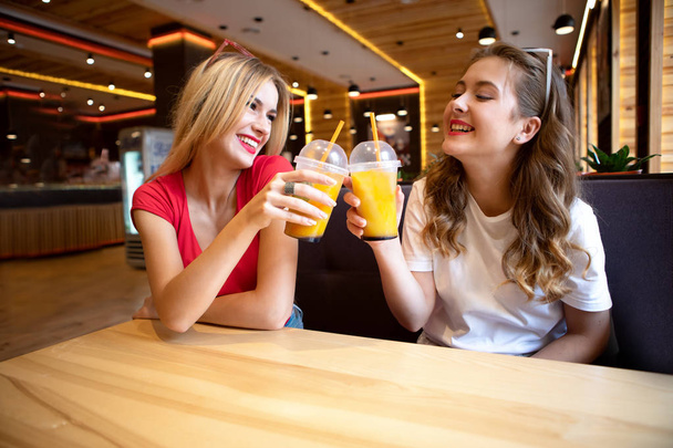 Kaksi ystävää juo cocktaileja kahvilassa. Nuoret hoikat opiskelijat lepäävät kahvilassa. Naura ja pidä hauskaa. tyylikkäästi pukeutunut lyhyisiin denim-shortseihin ja kirkkaisiin t-paitoihin
 - Valokuva, kuva