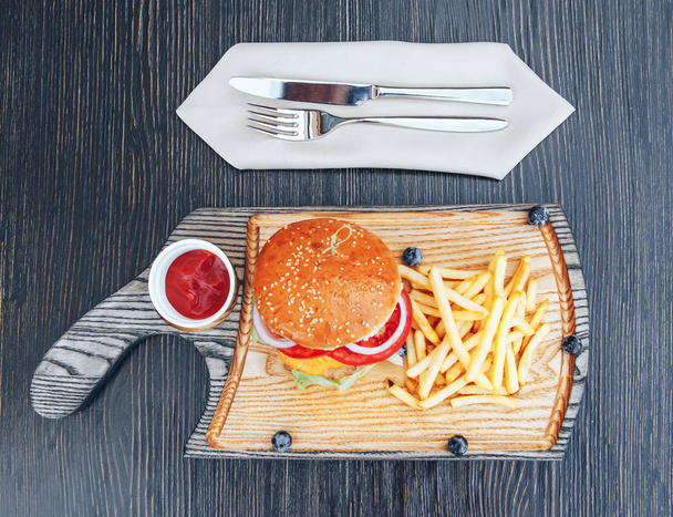 Fast food dish. Burger with fries on a wooden cutting board. Delicious hamburger and french fries with sauces. Cheeseburger with chicken meat - Photo, Image