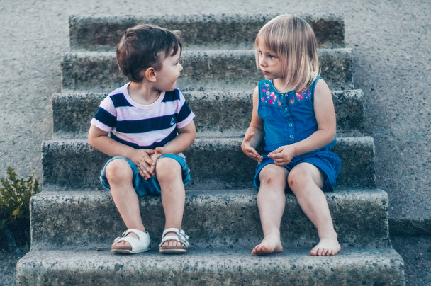 Два маленьких ребенка, девочка и мальчик на природе, сидят на камне, смотрят друг на друга с улыбкой, снаружи. Ретро тонизирован. Лето
 - Фото, изображение