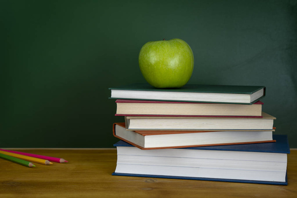Ένας δάσκαλος σε σχολείο της γραφείο με στοίβα από βιβλία ασκήσεων χρωματιστά μολύβια και πράσινο μήλο. Ένα πράσινο μαυροπίνακα - Φωτογραφία, εικόνα