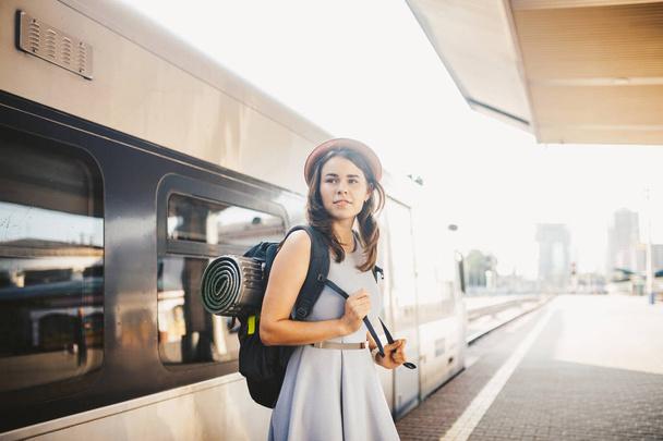 theme railway and travel. Portrait young caucasian woman with toothy smile standing at train station train background with backpack and equipment for tourism isolated mat in dress and hat in summer. - Foto, afbeelding