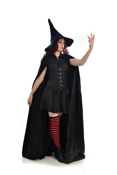 Ganzkörperporträt eines rothaarigen Mädchens, das einen langen schwarzen Mantel, einen spitzen Hut und ein Hexenkostüm trägt. Stehende Pose, isoliert auf weißem Studiohintergrund. - Foto, Bild
