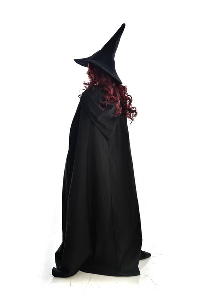 retrato de cuerpo entero de una chica pelirroja con una larga capa negra, sombrero puntiagudo y disfraz de bruja. pose de pie, aislado sobre fondo blanco del estudio
. - Foto, imagen