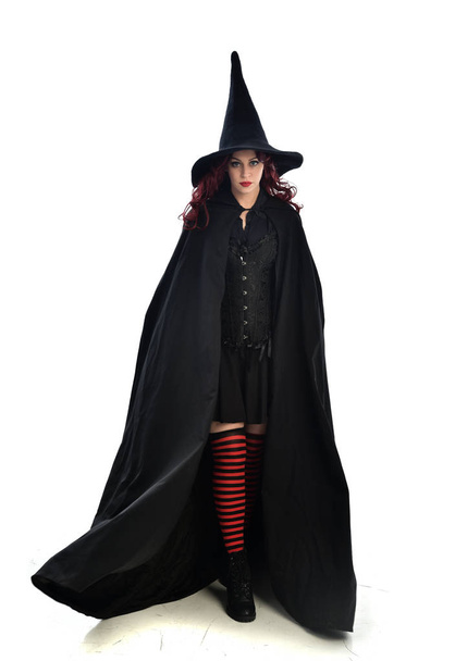 Pełna długość Portret rudowłosy dziewczyna sobie długi czarny płaszcz, kapelusz pointy i kostium czarownicy. Stojący poza, na białym tle na tle białego studia. - Zdjęcie, obraz