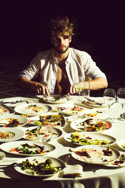 Handsome man or sexy muscular macho athlete in unbutton shirt eats with fork and knife at table with leftovers or residues food on dirty plates after banquet in restaurant on dark background. - Zdjęcie, obraz