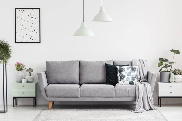 Grey sofa between cabinets with plants in white living room interior with lamps and poster. Real photo - Fotó, kép