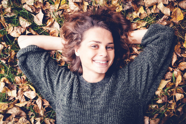 Beautiful young smiling girl woman with wavy hair lies on fallen autumn leaves, shot from above, portrait - 写真・画像