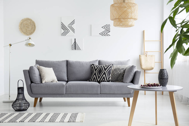 Real photo of a scandi living room interior with cushions on gray couch, cherries on wooden table and bag on a ladder - Фото, зображення