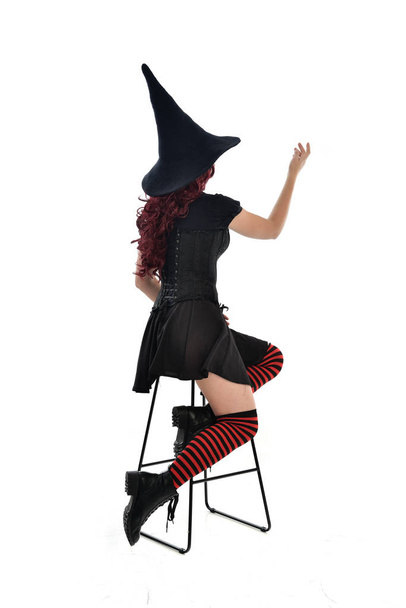 Ganzkörperporträt eines rothaarigen Mädchens, das einen langen schwarzen Mantel, einen spitzen Hut und ein Hexenkostüm trägt. sitzend auf Stuhl, isoliert auf weißem Studiohintergrund. - Foto, Bild