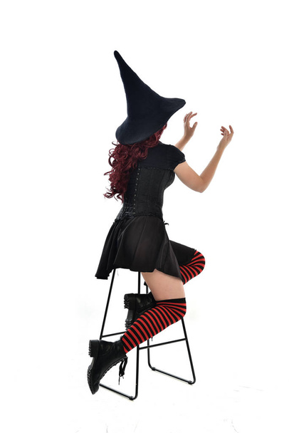 full length portrait of red haired girl wearing long black cloak, pointy hat and witch costume. seated pose on chair, isolated on white studio background. - Photo, Image