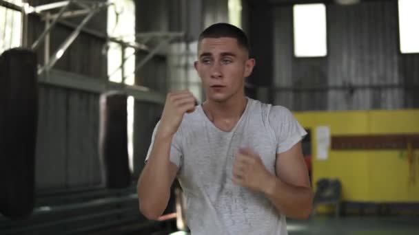 Front view footage of a training process. Young man boxing with invisible opponent, warming up. Male boxer in white T shirt working out in gym - Filmati, video