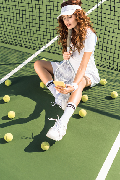 fashionable sportswoman in white sportswear with tennis racket taking selfie on smartphone at net on tennis court - Photo, Image