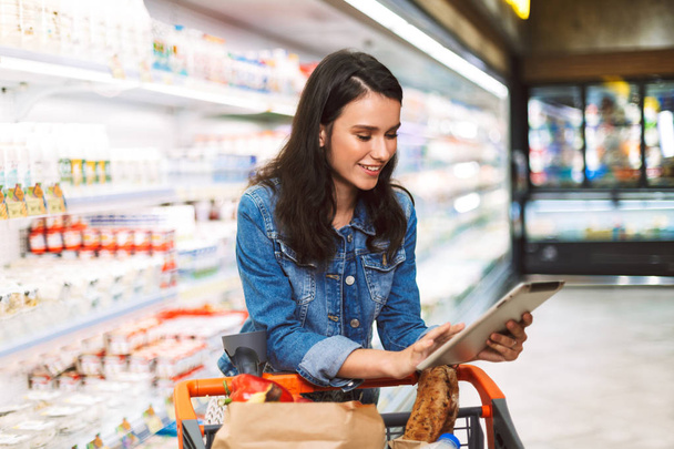 Souriante fille en jean veste avec chariot plein de produits heureux en utilisant la tablette dans le département des produits laitiers du supermarché moderne
 - Photo, image