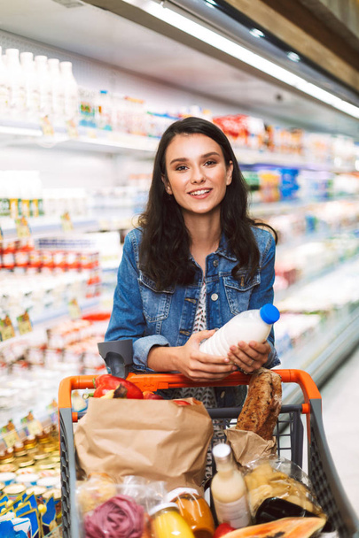 Hermosa chica sonriente en chaqueta de mezclilla con carro lleno de productos que miran soñadoramente en la cámara sosteniendo la botella de leche en las manos en el departamento de lácteos del supermercado moderno
 - Foto, imagen
