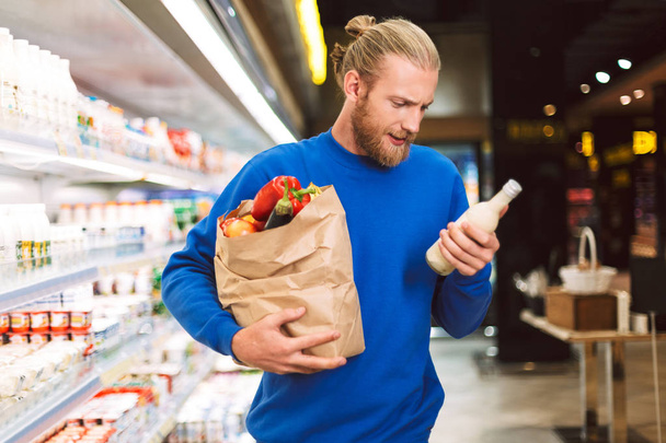 Młody człowiek w ciemny niebieski sweter trzymając papier spożywczy worek pełen produktów starannie wybierając mleko mleczne w departamencie nowoczesny supermarket - Zdjęcie, obraz