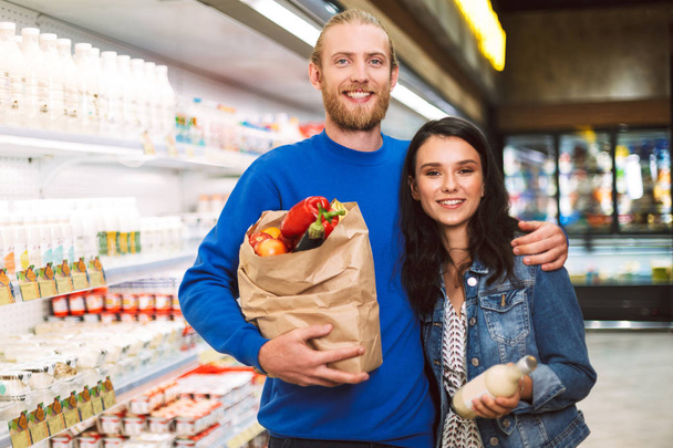 紙食料品と若い美しいカップル バッグ喜んでカメラを探して近代的なスーパー マーケットの乳製品部門の製品の完全な - 写真・画像