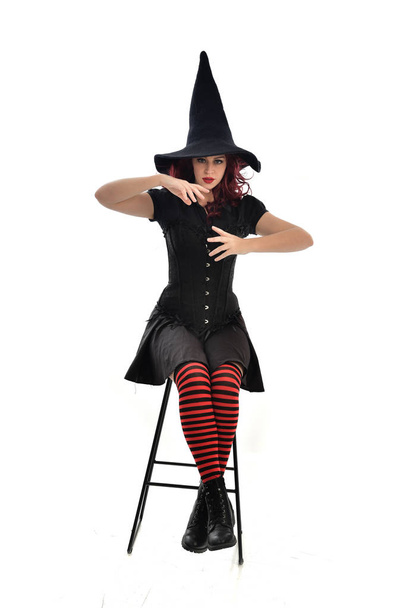 retrato de comprimento total da menina de cabelos vermelhos vestindo manto preto longo, chapéu pontiagudo e traje de bruxa. postura sentada na cadeira, isolada no fundo do estúdio branco
. - Foto, Imagem