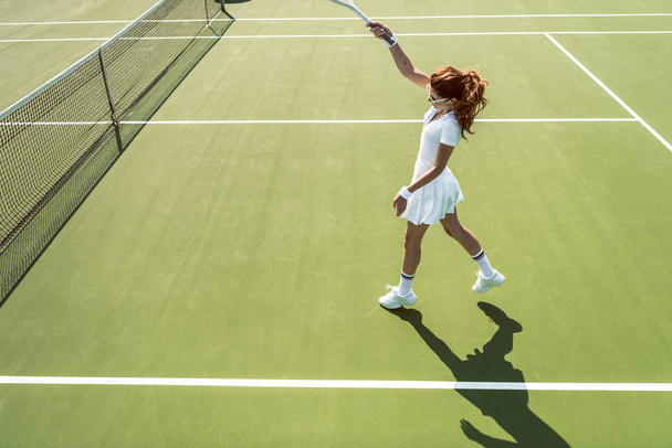 vista lateral de la joven atractiva mujer en uniforme de tenis blanco jugando tenis en la cancha
 - Foto, imagen