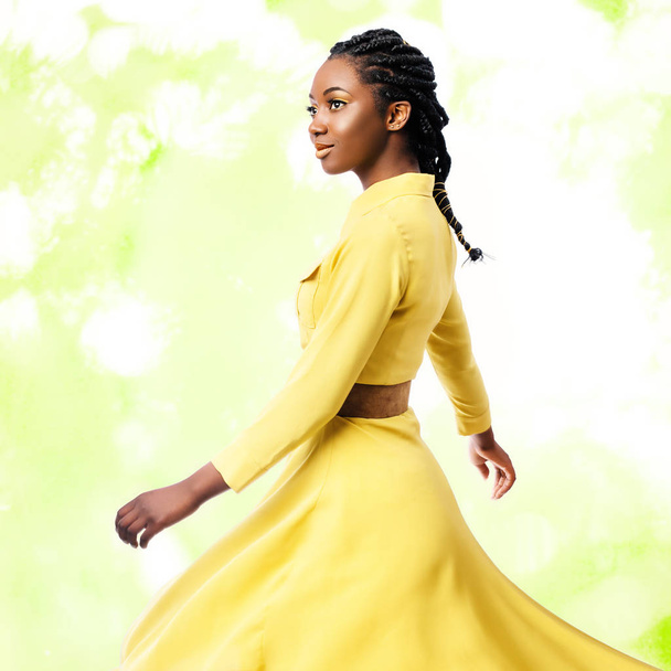 緑背景にカラフルな絹のような黄色のドレスを揺れる編みこみの髪型を持つ若い魅力的なアフリカ女性のスタジオ ポートレート  - 写真・画像