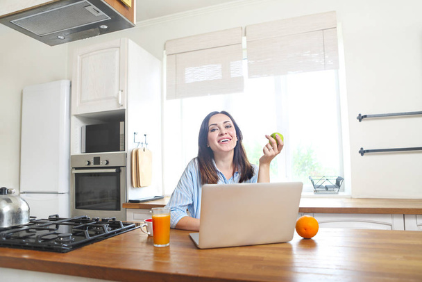 Νεαρή γυναίκα με τον υπολογιστή και το φλιτζάνι του καφέ στην κουζίνα το πρωί - Φωτογραφία, εικόνα