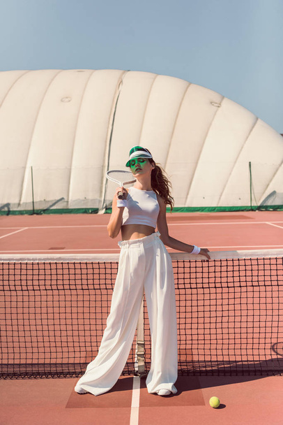 fashionable woman in white clothing and cap with tennis racket posing at tennis net on court - Foto, imagen