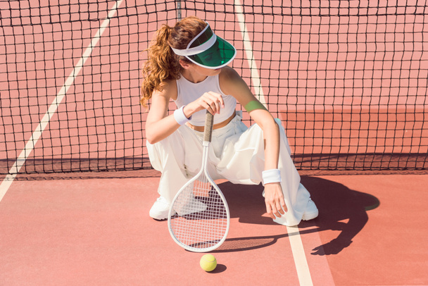 fashionable woman in white clothing and cap with tennis racket sitting at tennis net on court - Photo, Image