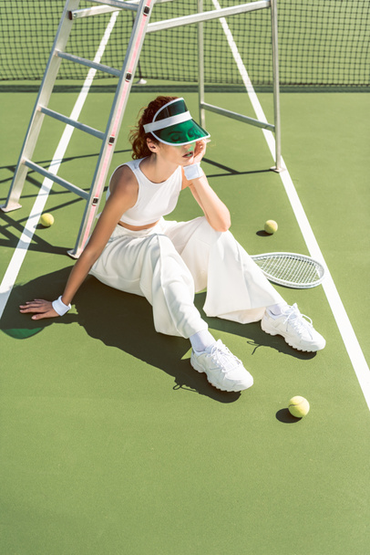 νεαρή γυναίκα σε μοντέρνα λευκά ρούχα και καπάκι κάθεται στο γήπεδο τένις με ρακέτα και μπαλάκια - Φωτογραφία, εικόνα