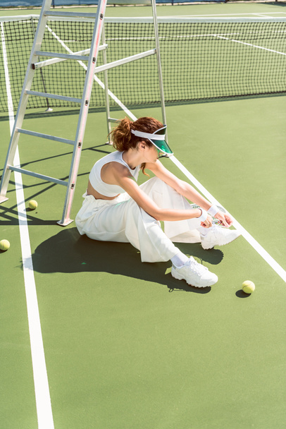 вид збоку молодої жінки в стильному білому одязі і кепці зав'язують шнурки на тенісному корті з ракеткою і кульками
 - Фото, зображення