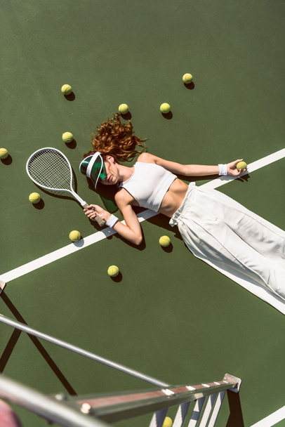 overhead view of stylish woman in white clothing and cap lying with racket lying on tennis court with racket - 写真・画像