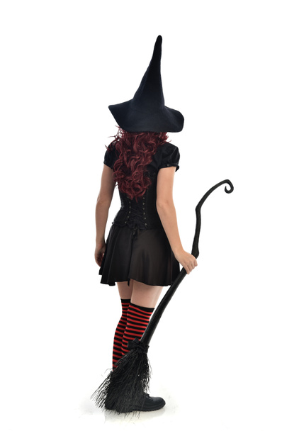 полноразмерный портрет рыжеволосой девушки в чёрном костюме ведьмы и остроконечной шляпе, держащей метлу. Стоя поза, изолированные на белом фоне студии
. - Фото, изображение