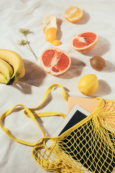 tablet computer in yellow string bag and fresh ripe tropical fruits   - Fotografie, Obrázek