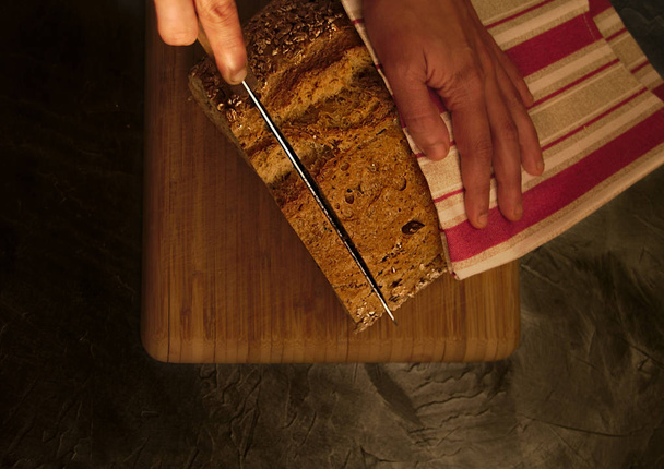 Overhead picture of natural bread with seeds, over wood table, black background, and hand with knife cutting a slide of bread, including red and brown cloth - Фото, изображение